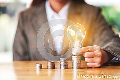 Businesswoman holding and putting light bulb on coins stack on the table for saving energy and money Stock Photo