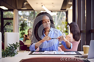 Businesswoman Sitting At Desk Taking Photo Of Proofs Or Design Layouts On Mobile Phone Stock Photo