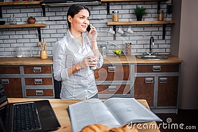 Businesswoman stand in kitchen and talk on phone. She hold white cup with drink. Laptop jjournal and plate with Stock Photo