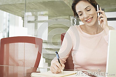 Businesswoman Using Mobile Phone While Writing On Notepad Stock Photo