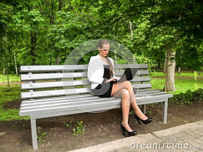 Businesswoman working on laptop sitting on a bench in the park. Cute young adult girl in a little black dress, white jacket and Stock Photo