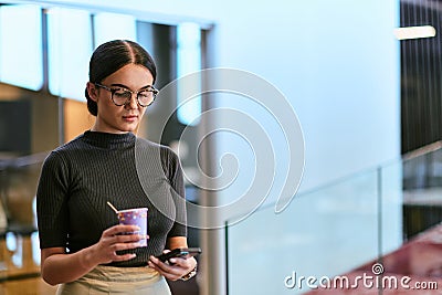 In a bustling modern office a businesswoman in glasses juggles her tasks, sipping coffee and using her smartphone Stock Photo