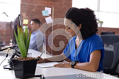 Busy african american businesswoman sitting at desk with hygiene screen writing in office Stock Photo