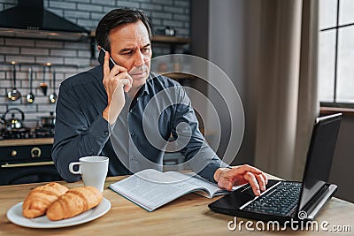 Busy man sit at table in kitchen and talk on phone. He type on keyboard laptop and work. Journal white cup and plate Stock Photo