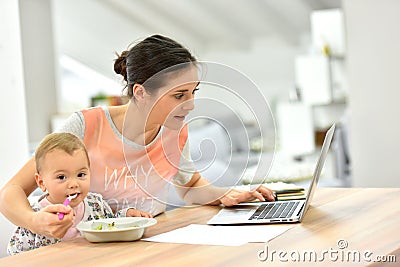 Busy mother working on laptop and feeding her baby Stock Photo