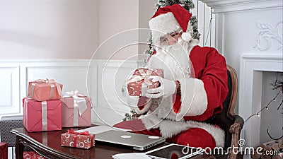 Busy Santa Claus preparing presents using laptop and digital tablet, sorting his letters and receiving a gift from a Stock Photo