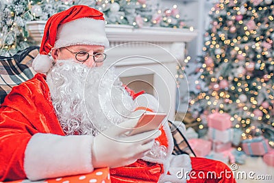Busy Santa Claus sits and look at phone he holds in hands. Man is alone in festive room. He sits at fireplace and Stock Photo