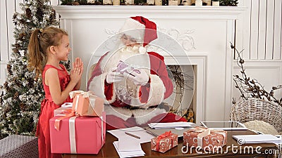 Busy Santa sorting chidlren`s letters while cute little helper bringing him a gift Stock Photo