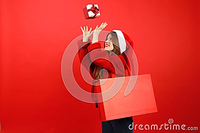 Buy gifts for Christmas, sale of New Year's goods. Cheerful young woman in santa hat throws up a box on a red background Stock Photo