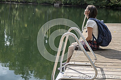 A calm, dreaming woman with a backpack sits on a wooden pier by the lake, in the rays of the warm summer sun Stock Photo