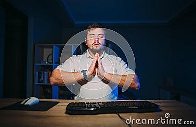 A calm man meditates at night on the computer, closes his eyes, raises his hands and tries to calm down Stock Photo