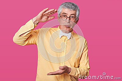Calm senior shows large or big object, stands against pink wall, dressed in stylish yellow shirt and white bowltie, has serious Stock Photo