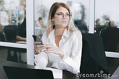 Calm serious young attractive blonde uses phone while working at table in office Stock Photo