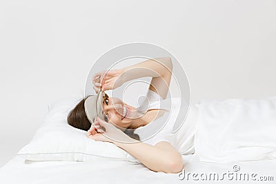 Calm young brunette woman lying in bed with sleep mask on white sheet, pillow, blanket on white background. Smiling Stock Photo