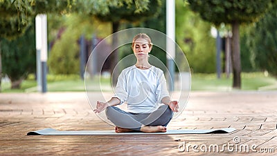 Calm young woman sitting in lotus pose on mat during morning meditation in park Stock Photo