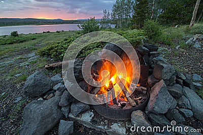 A campfire burns hot at a lakeshore campsite, as the sun sets in the mountains. Stock Photo