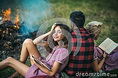 Camping in wilderness. Turn back guy looking at fire while two beautiful girls read book. Smiling brunette with braces Stock Photo