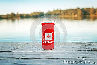 Can of beer in red cozy beer can cooler with Canadian flag standing on wooden pier by lake outdoor. Celebrating Canada Day Stock Photo