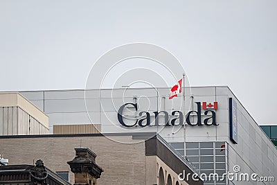 Canada Wordmark, the official logo of the Canadian government, on an administrative building next to a Canadian flag waiving Editorial Stock Photo