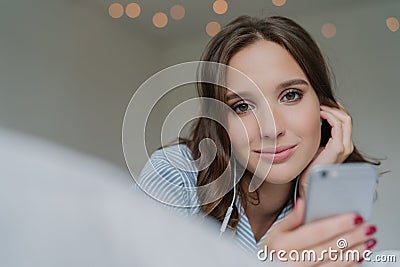 Candid shot of pretty dark haired woman spneds free time with favourite song, holds modern cell phone, looks directly at camera, p Stock Photo