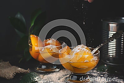 Canned peaches with syrup in a bowl on a dark background, near a tin can with a spoon, powdered sugar sprinkling on top, motion in Stock Photo