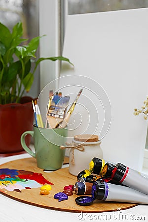 Canvas, wooden artist's palette with colorful paints and brushes on white table near window Stock Photo