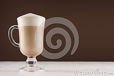 Cappuccino coffee in elegant glass with foam on white table and dark brown wall, copy space. Stock Photo