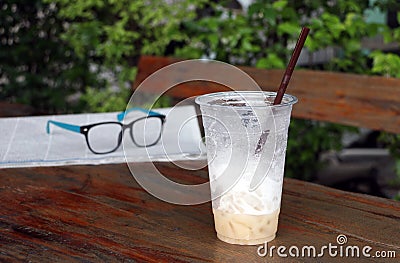 Less cappuccino coffee in the plastic cup and out focus spectacles on the newspaper on the wooden table. Stock Photo