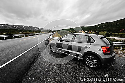 Grey modern car is parking next to a rural paved road which leads through the nature of Norway as far as the eye can see Stock Photo