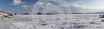 Car tracks lead from the shore to a snowy frozen large lake with forest mountains on the horizon in the winter sunny day Stock Photo