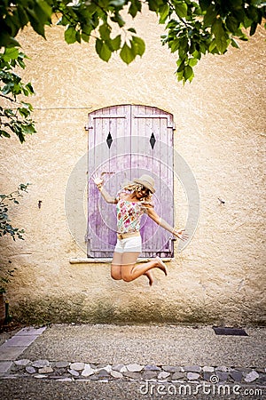 Carefree beautiful woman in hat jumping midair in front of old wooden closed window of textured building wall. Ecstatic hipster Stock Photo