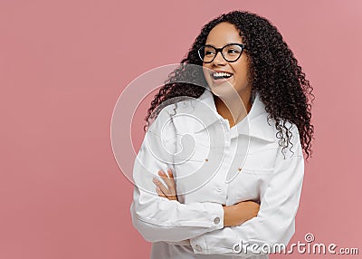Carefree cheerful Afro American woman wears white jacket, keeps arm folded, looks aside, has charming smile, enjoys nice day in Stock Photo