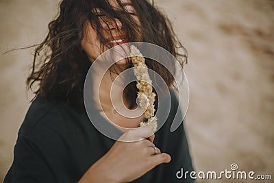 Carefree hipster girl with windy hair sitting and smiling on sandy beach, holding herb. Stylish tanned young woman in modern Stock Photo