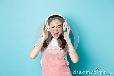Carefree pretty girl scream from joy, listening music in headphones with happy face, lip-sync lyrics, standing over blue Stock Photo