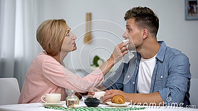 Careful mother wiping adult sons mouth during having tea at home, overprotection Stock Photo