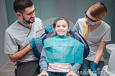 Careful positive male and female dentist look at kid patient and smile. Girl sit in dental chair. She look straight and Stock Photo