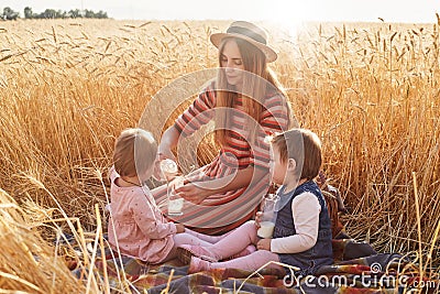Caring helpful mother sitting on blanket at wheat field with her lottle daughters, filling glasses with milk from bottle, wearing Stock Photo