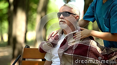 Caring nurse covering blind pensioner with blanket outdoors, home for elderly Stock Photo