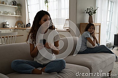 Caring puzzled mother sits on couch with phone and looks back at offended daughter after punishment Stock Photo