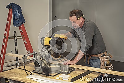 Carpenter with tool belt cutting wood boards with electric chop saw at saw horses during home remodel construction Stock Photo