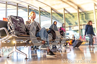 Casual blond young woman using her cell phone while waiting to board a plane at bussy airport departure gates. Stock Photo