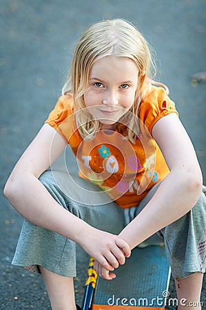 Casual, pretty blonde girl with enigmatic smile sitting on a skateboard and making eye contact Stock Photo