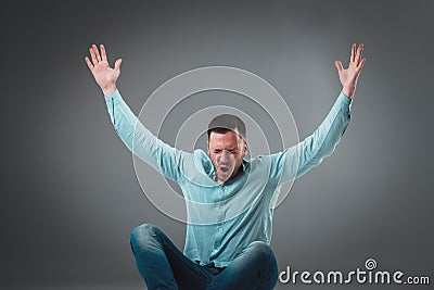 Casual young man sitting on the floor with his legs crossed and cheering with his hands in the air while screaming. Stock Photo