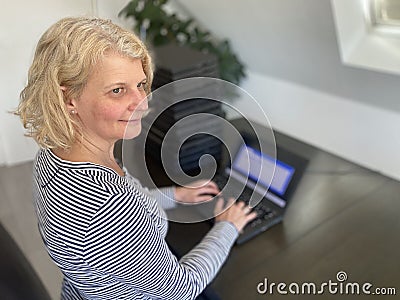 A casually dressed caucasian middle aged woman is installing a laptop computer with a stack of several more computers Stock Photo