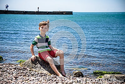 Caucasian baby boy is sitting at the beach. Cute child is playing with roks and shellfishes muscle-fish on the sand in summer day Stock Photo
