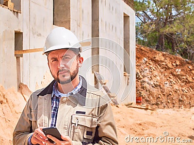 Caucasian bearded civil engineer or construction worker in white hardhat uses his pnone opposite construction site Stock Photo