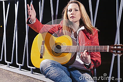 Caucasian Blond woman Posing in Red Leather Jacket and Jeans with Guitar Outdors on Dark Street Stock Photo