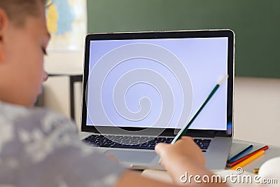 Caucasian boy sitting at a desk in classroom writing and using laptop, with copy space on screen Stock Photo