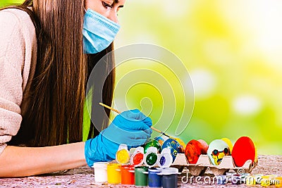 Caucasian concentrated girl in medical mask and disposable gloves decorates handmade eggs for the pascal holidays in Stock Photo