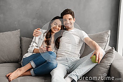 Caucasian couple man and woman in casual clothing sitting on sofa in apartment, while having breakfast with apple and tea Stock Photo